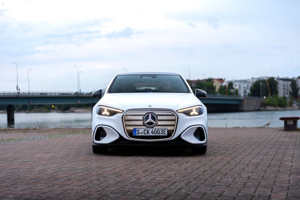 A white Mercedes-Benz C-Class Electric seen from the front, parked on a brick waterfront area with a bridge and river in the background.