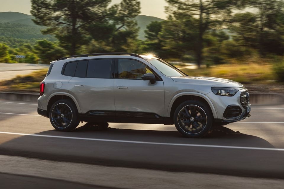 A silver Mercedes-Benz GLB SUV driving along a road, seen from the side, with a blurred tree and mountain backdrop