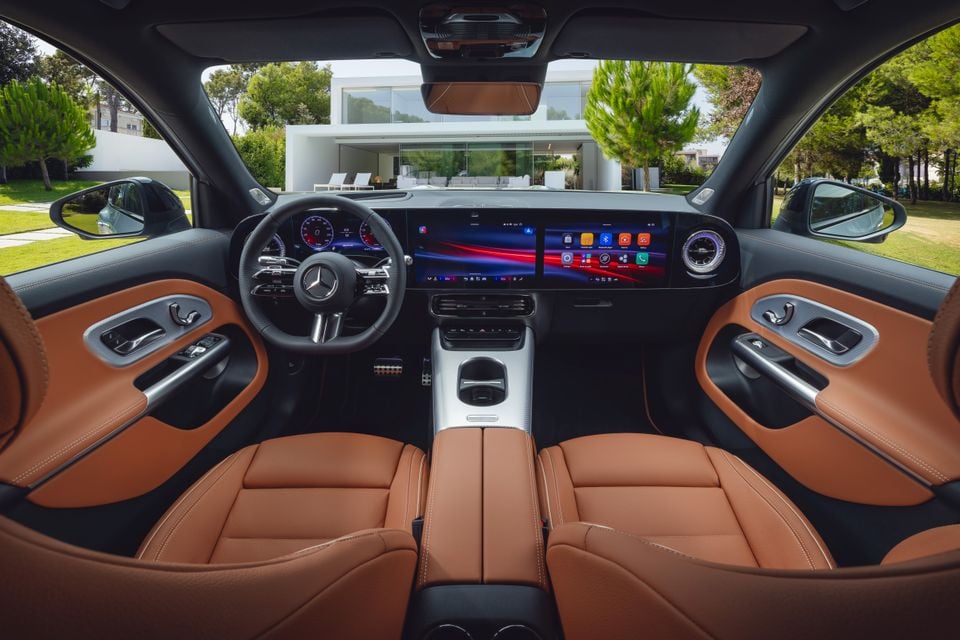 Interior of a 2027 Mercedes-Benz GLB showing tan leather front seats, a wide dual-screen infotainment and instrument display, and a flat-bottomed AMG steering wheel