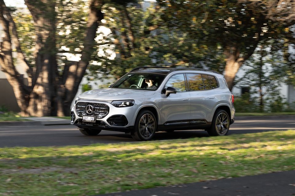 A silver 2027 Mercedes-Benz GLB 350 4Matic electric SUV driving along a tree-lined street, seen from the front left