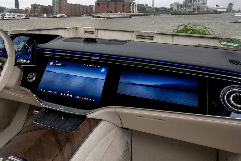 The dashboard of a Mercedes-Benz S-Class shows dual widescreen infotainment displays with a river and city skyline visible through the windscreen.