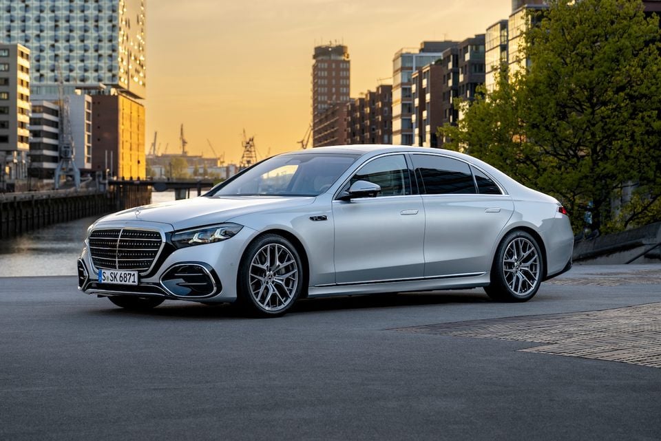 A silver Mercedes-Benz S-Class sedan parked near a waterfront with city buildings in the background at sunset.