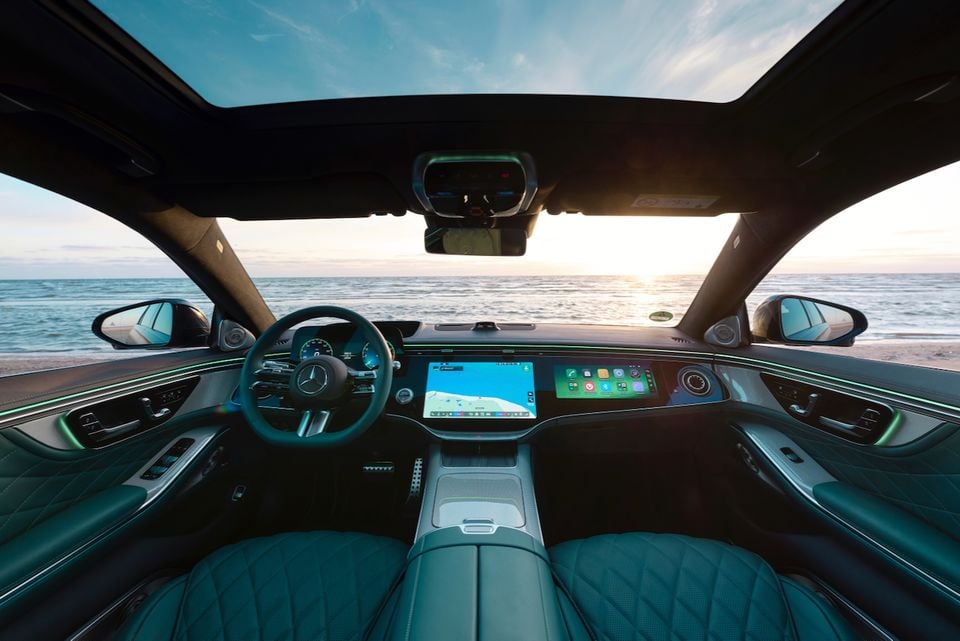 The interior of a 2027 Mercedes-Benz S-Class shows the steering wheel, dashboard screens and teal leather seats with a beach visible through the windscreen.