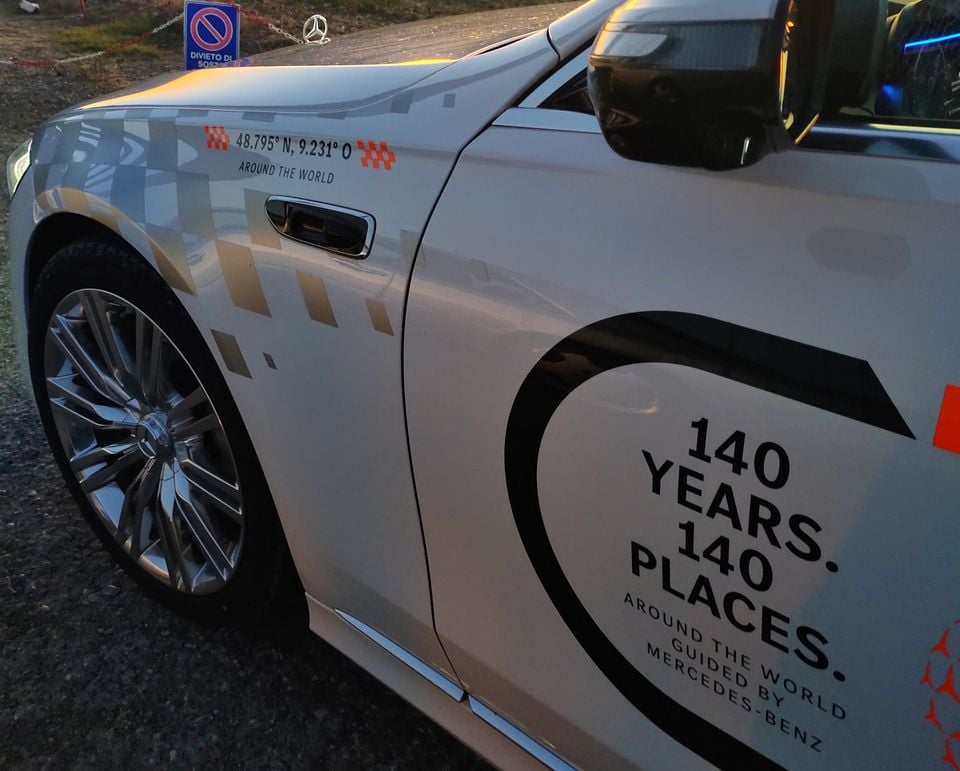 Close-up side view of a white Mercedes-Benz S-Class wearing "140 Years. 140 Places." promotional livery on its front door and fender