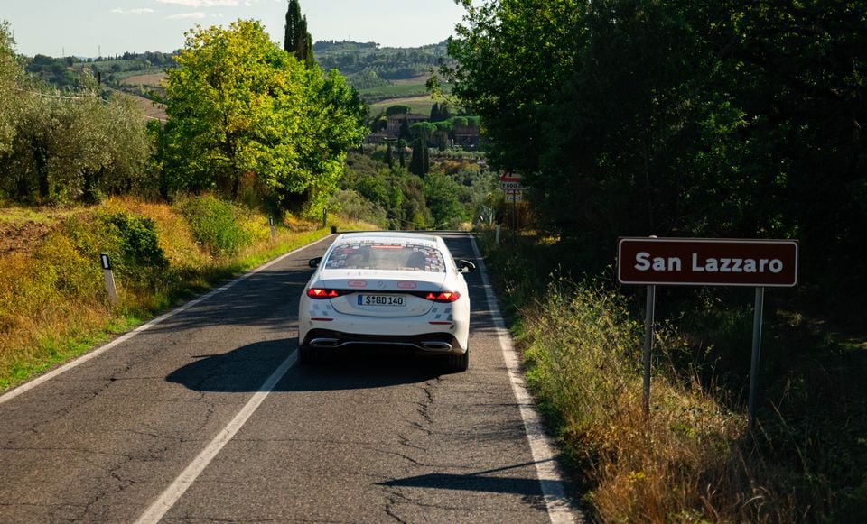 A white Mercedes-Benz S580L sedan, covered in camouflage decals, driving away on a rural Italian road past a San Lazzaro town sign.