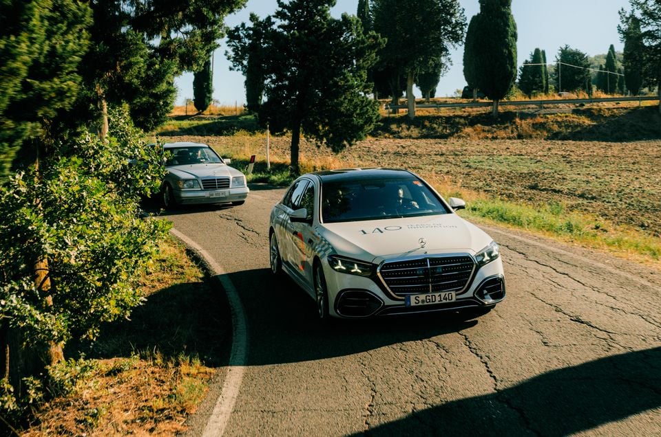 A white Mercedes-Benz S-Class sedan drives on a rural road with an older Mercedes-Benz sedan following behind on a sunny day