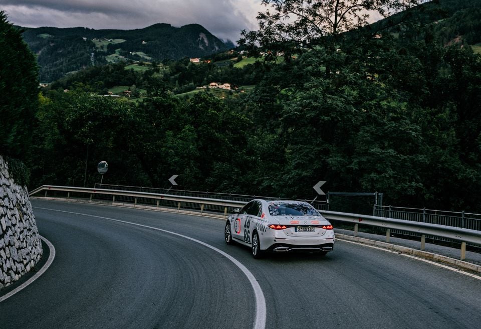 A white 2027 Mercedes-Benz S580L prototype sedan, covered in camouflage decals, driving on a mountain road, seen from the rear