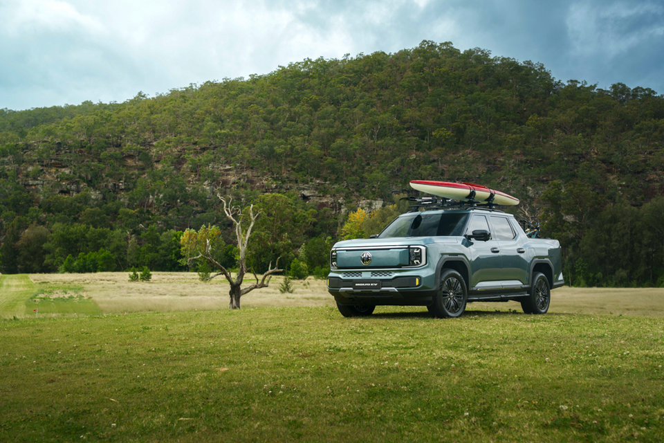 A green-grey 2027 MG U9 EV dual-cab ute with a red kayak on its roof racks parked on a grassy field below a forested hill
