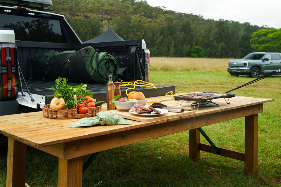 A 2027 MG U9 electric ute parked in a grassy field with its tailgate open, powering a barbecue grill on a picnic table via an extension cord