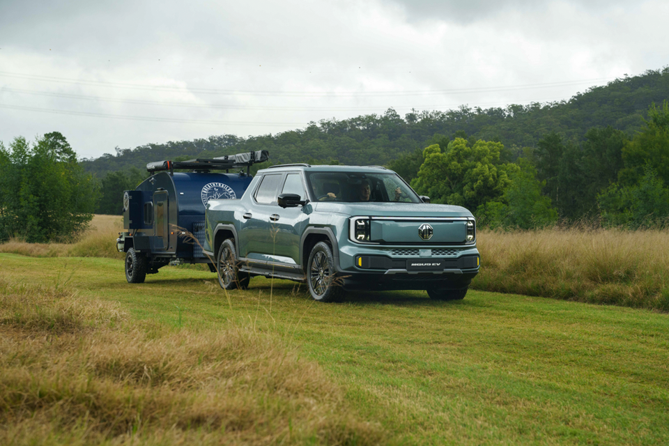 A green-grey 2027 MG U9 EV dual-cab ute towing a blue off-road camper trailer across a grassy paddock, seen from the front