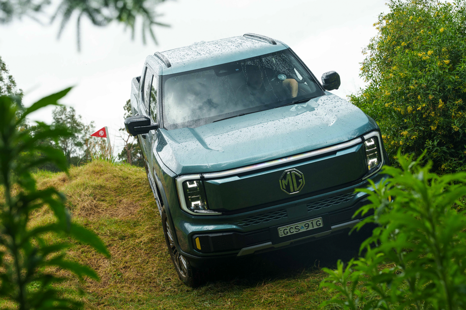 A teal 2027 MG U9 electric dual-cab ute driving down a grassy mound on an off-road course, seen from the front