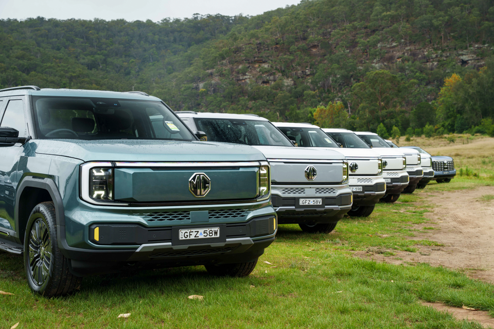 A row of 2027 MG U9 electric utes parked on grass beside a dirt track and bushy hillside, led by a green-grey example with white ones behind