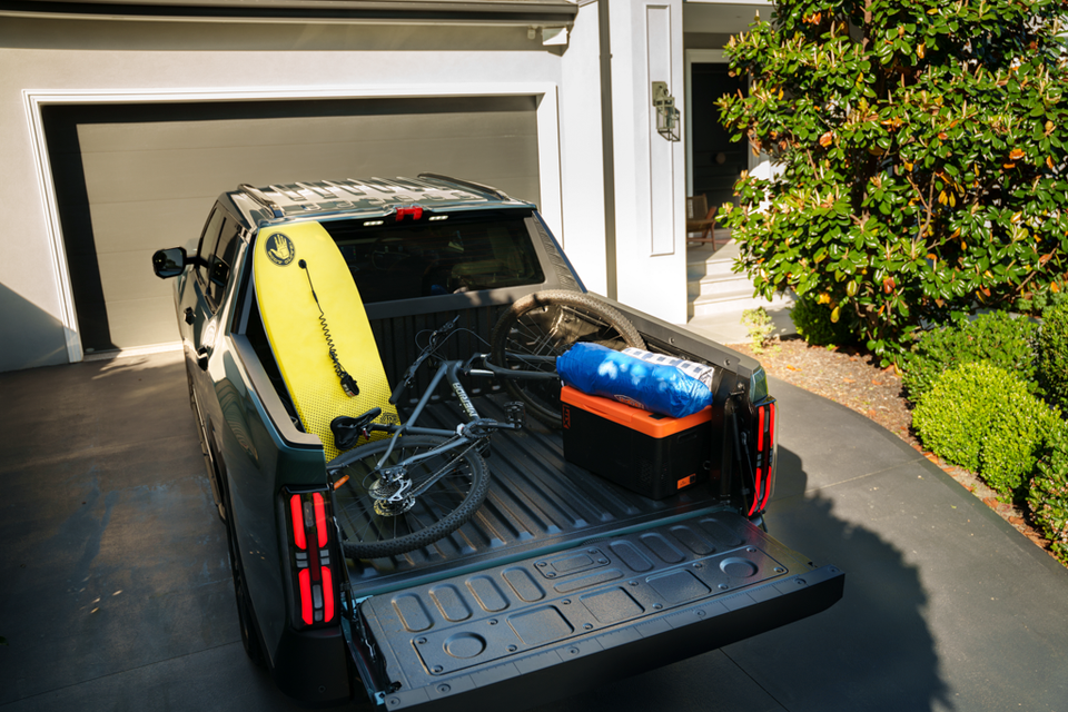 Dark green 2027 MG U9 EV ute parked in a driveway, seen from the rear with the tailgate open and a bodyboard, bike and cooler box in the tray