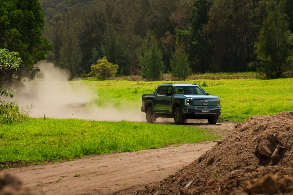 A green 2027 MG U9 EV dual-cab ute driving along a dirt track through a grassy paddock, kicking up a cloud of dust