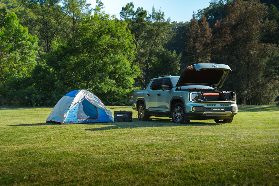 A green 2027 MG U9 EV dual-cab ute parked on grass with its front trunk open, beside a camping tent and a storage box in bushland