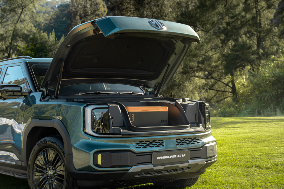 A teal 2027 MG U9 EV ute parked on grass with its bonnet raised, revealing a front storage compartment holding a cooler box