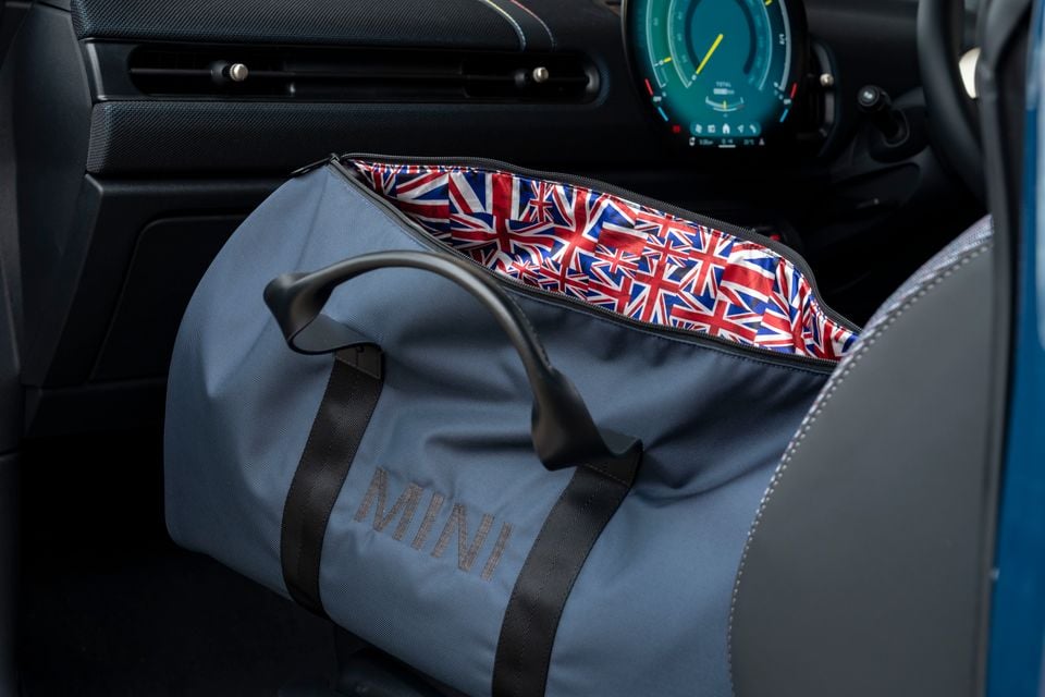 A MINI-branded grey duffle bag with Union Jack-patterned lining sitting on the passenger seat of a Mini Cooper, with the digital instrument cluster visible in the background
