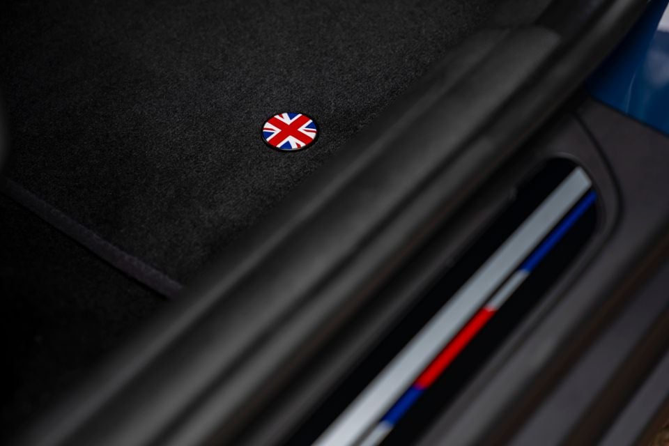 Close-up of a Union Jack badge on a dark carpet floor mat inside a Mini Cooper Oxford Edition, with a coloured sill plate visible nearby