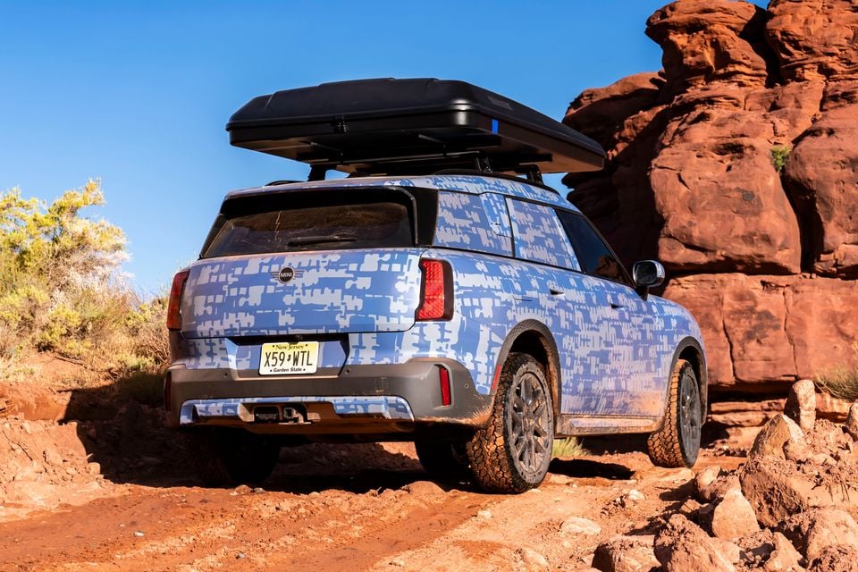 A camouflaged MINI Countryman SUV seen from the rear, navigating a rocky off-road trail with red rock formations in the background and a roof box mounted on top