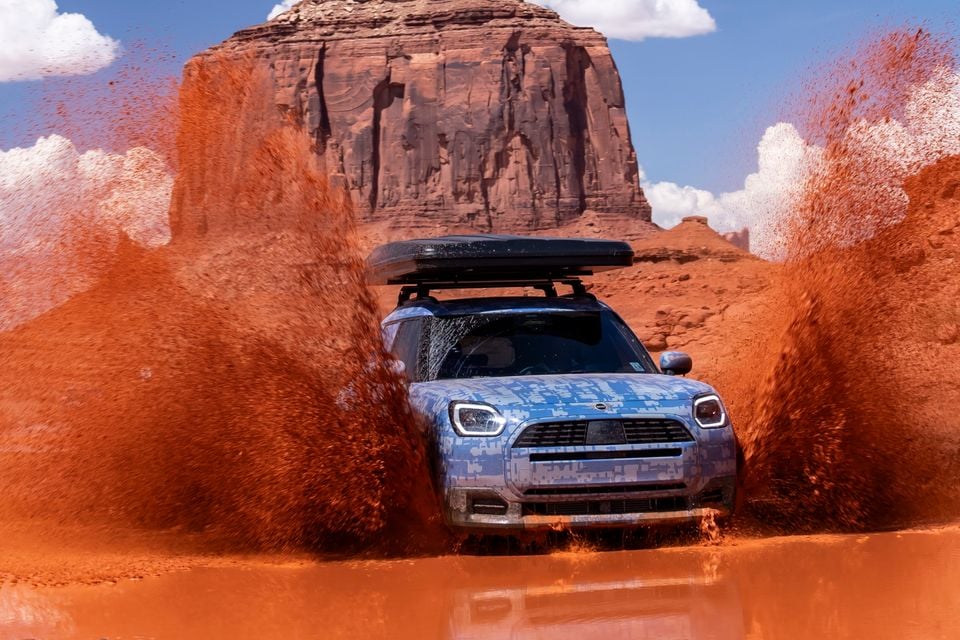 A camouflaged MINI Countryman SUV with a roof box drives through red mud in a desert landscape, sending large sprays of red dirt into the air