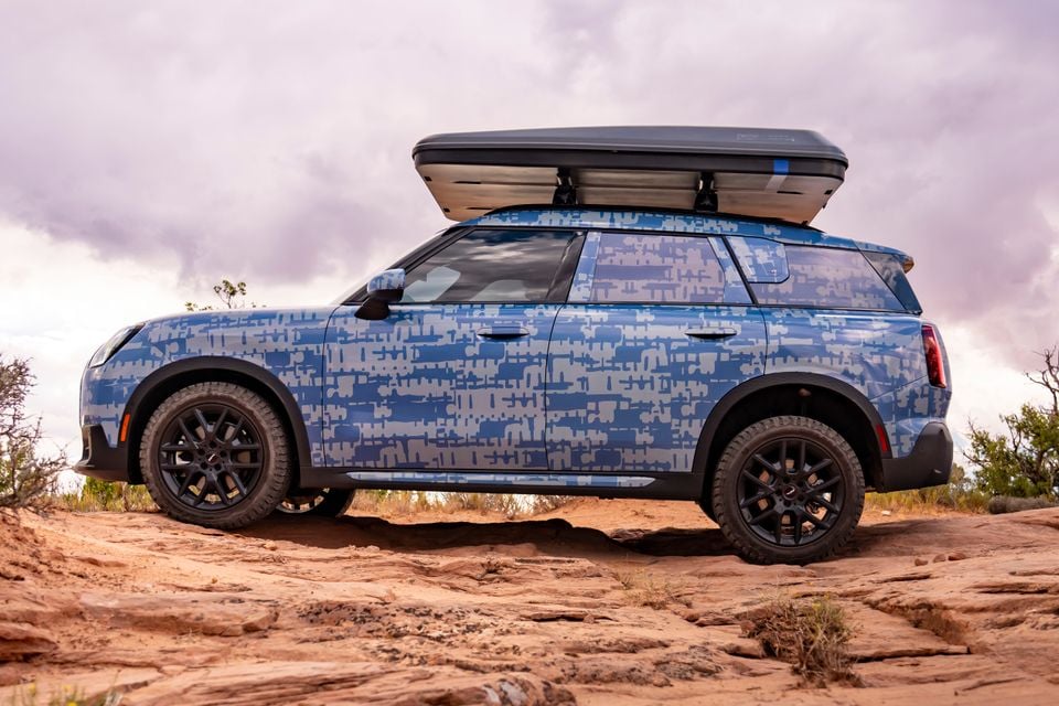 A MINI Countryman SUV with a blue camouflage wrap and roof cargo box, seen from the side on rocky terrain outdoors