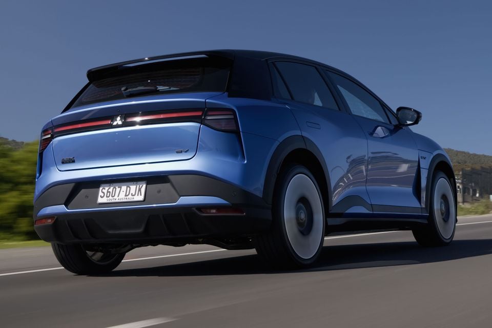A blue 2027 Mitsubishi ASX EV small SUV driving on a road, seen from the rear quarter.