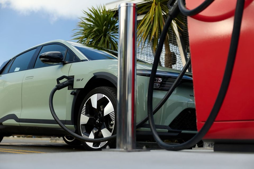 A green Mitsubishi ASX electric SUV plugged into a charging cable at an EV charging station, with a palm tree in the background.