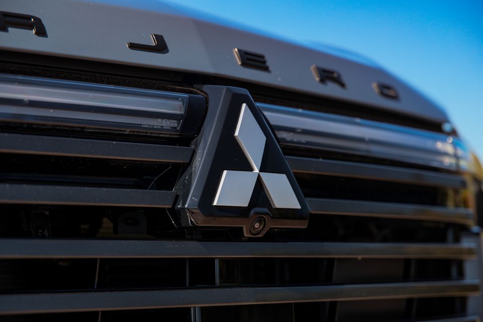 Close-up of the Mitsubishi three-diamond badge with front camera on the dark grille of a 2027 Mitsubishi Pajero, with PAJERO lettering on the bonnet