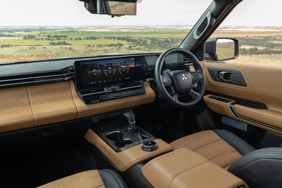 Dashboard of the 2027 Mitsubishi Pajero with tan leather trim, a wide widescreen display, steering wheel and centre console, overlooking farmland