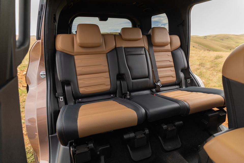 Rear bench seats in tan and black leather inside a 2027 Mitsubishi Pajero, seen through the open rear door with grassy hills outside