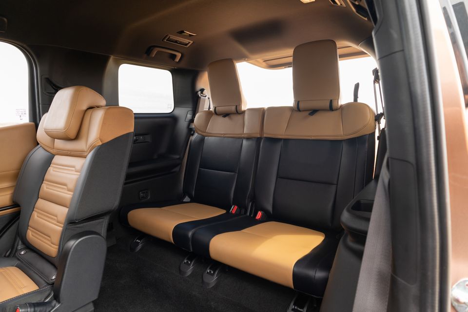 Third-row bench seats in tan and black leather inside a 2027 Mitsubishi Pajero, seen through the open rear door beside a second-row seat