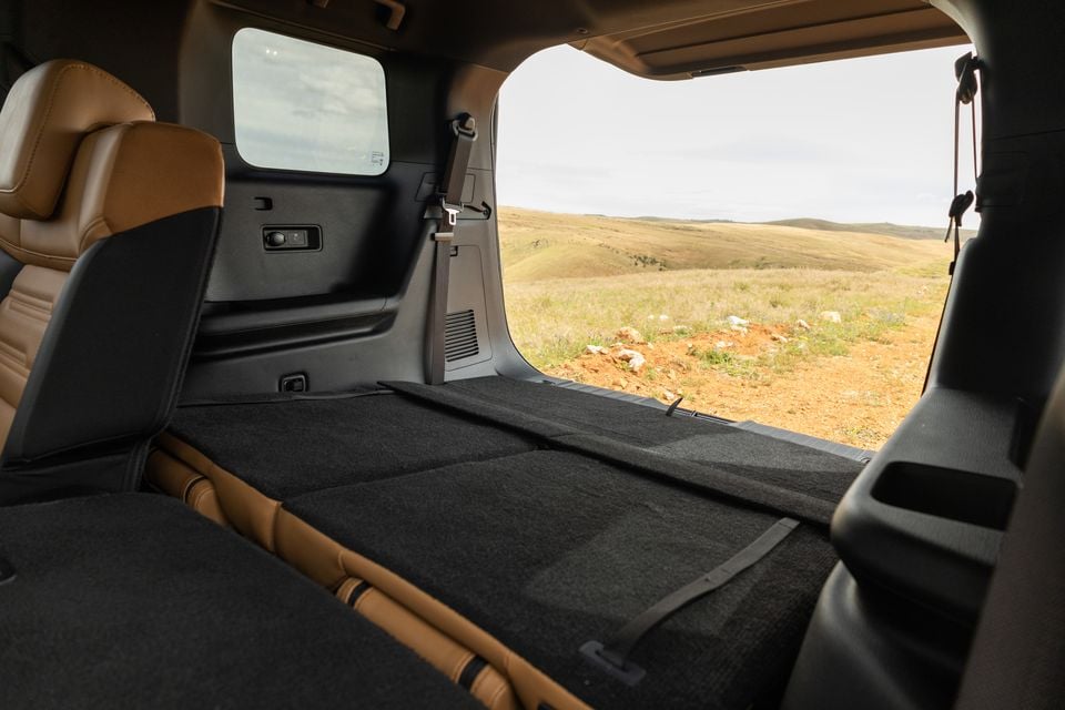 Folded third-row seats forming a flat cargo floor inside a 2027 Mitsubishi Pajero, with the tailgate open to grassy hills