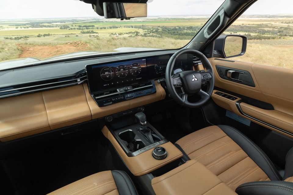 Interior of the 2027 Mitsubishi Pajero showing a tan leather dashboard, widescreen digital displays, steering wheel and front seats overlooking farmland