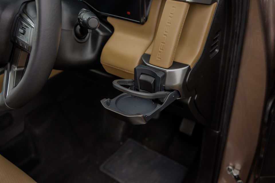 Fold-out cupholder below the dashboard of a 2027 Mitsubishi Pajero, beside tan leather trim embossed with the Pajero name and the steering wheel