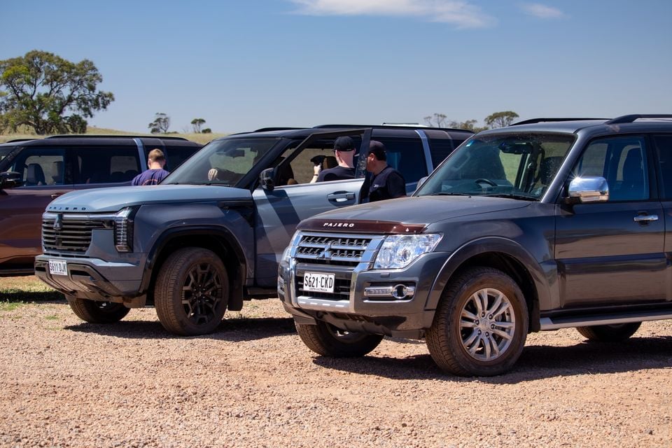 The new 2027 Mitsubishi Pajero parked beside the previous-generation Mitsubishi Pajero on gravel, seen from the front, with people standing nearby