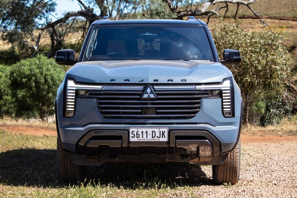 A grey-blue 2027 Mitsubishi Pajero SUV seen from the front, parked on gravel among native bushland