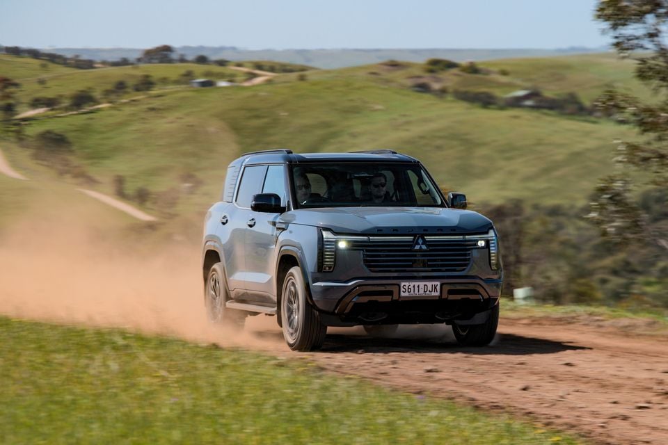 A grey 2027 Mitsubishi Pajero SUV driving along a dusty dirt track through green rolling hills, seen from the front.