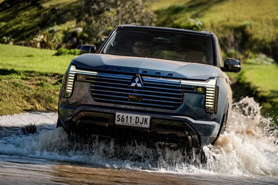 A grey 2027 Mitsubishi Pajero SUV driving through a shallow water crossing, splashing water, seen from the front