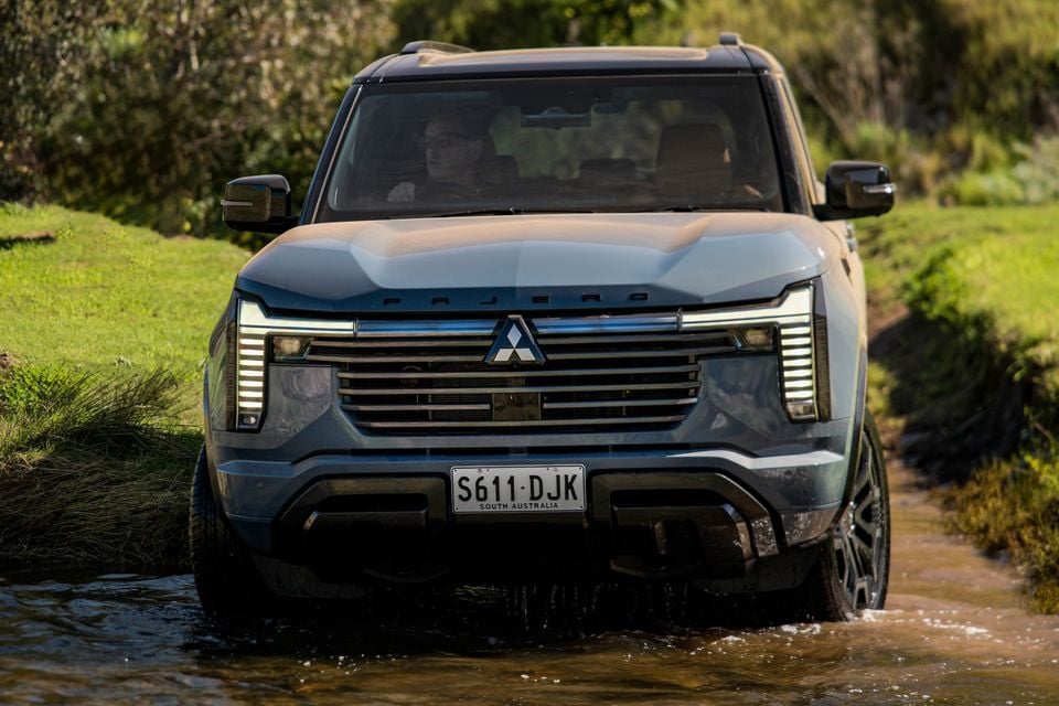 A grey 2027 Mitsubishi Pajero SUV driving through a shallow creek crossing, seen from the front