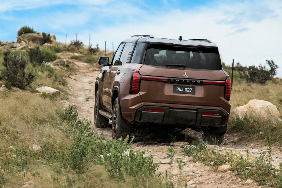 A bronze 2027 Mitsubishi Pajero SUV seen from the rear, driving up a rocky off-road track through grassy hills