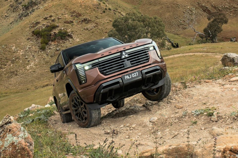A bronze 2027 Mitsubishi Pajero SUV climbing a rocky dirt track off-road in grassy hill country, seen from the front