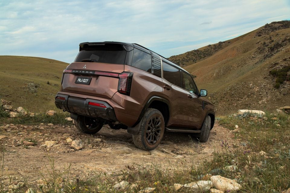 A brown 2027 Mitsubishi Pajero SUV driving up a rocky dirt track between grassy hills, seen from the rear