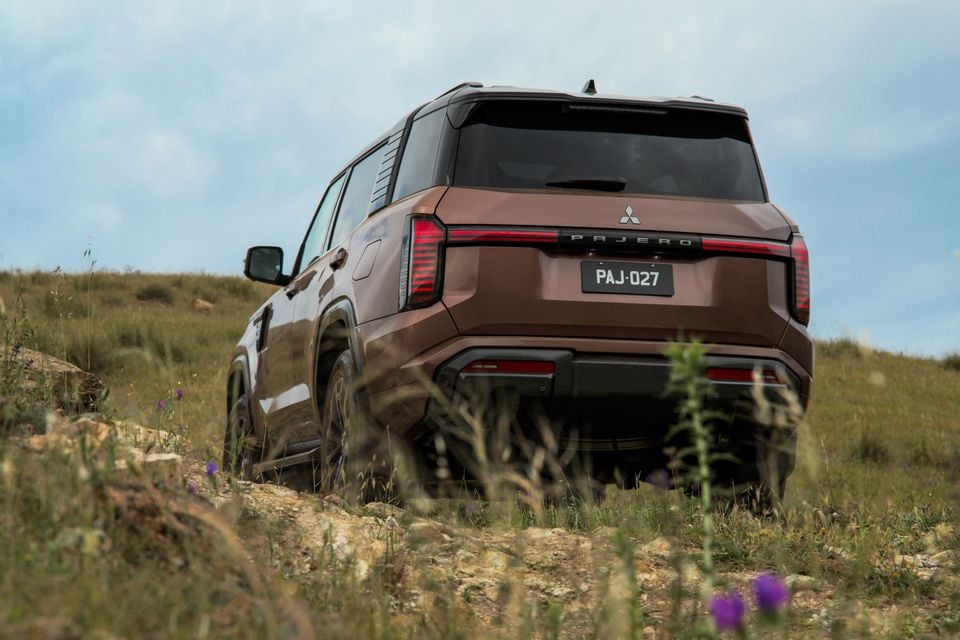 A bronze 2027 Mitsubishi Pajero SUV climbing a rocky grassy hill off-road, seen from the rear