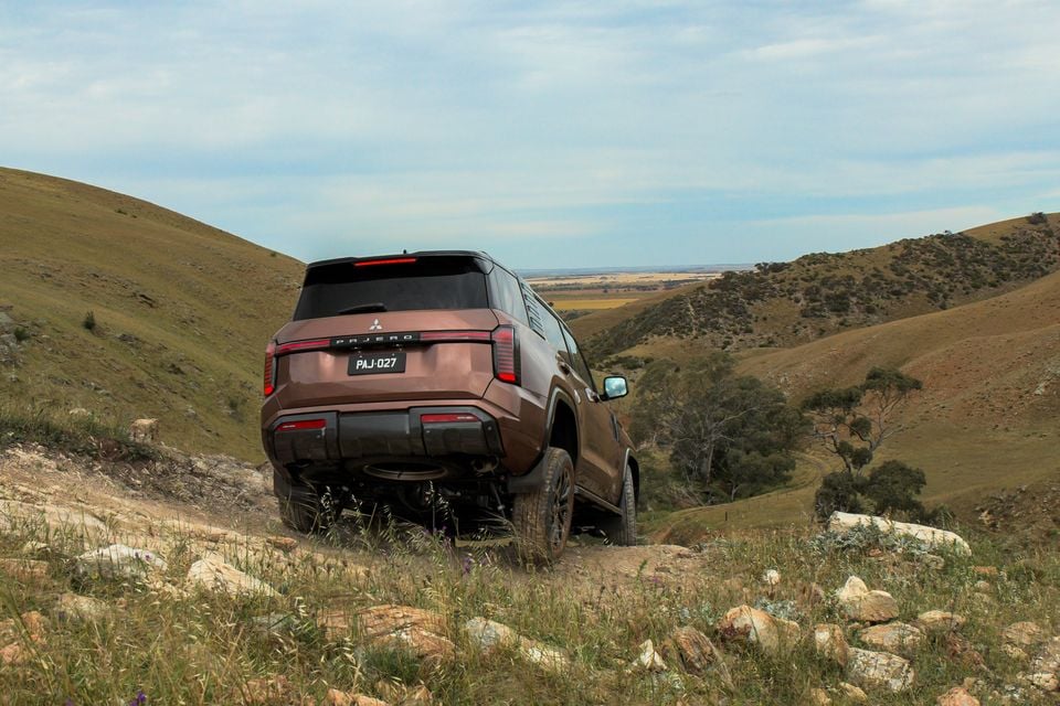 A bronze 2027 Mitsubishi Pajero SUV seen from the rear, driving down a rocky off-road track through grassy hills
