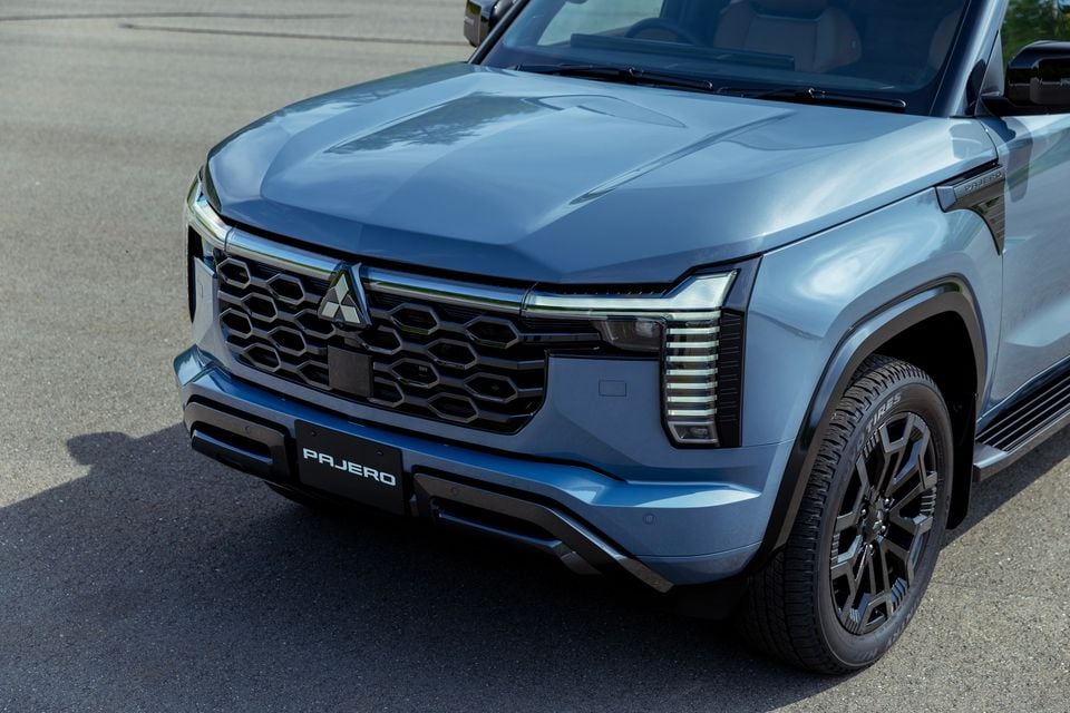 Close-up of the front end of a blue Mitsubishi Pajero SUV, showing the bold hexagonal grille, Mitsubishi logo, layered headlights, and black alloy wheel.