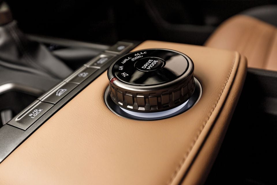 Close-up of the drive mode selector dial and 4WD transfer case knob on the centre console of a 2027 Mitsubishi Pajero, showing 2H, 4H, 4HLc and 4LLc settings, set into tan leather trim with additional off-road control buttons nearby.
