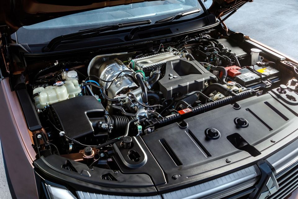 The engine bay of a Mitsubishi Pajero with the bonnet open, showing the turbocharged engine and components including the battery, coolant reservoir, and Mitsubishi-branded engine cover.