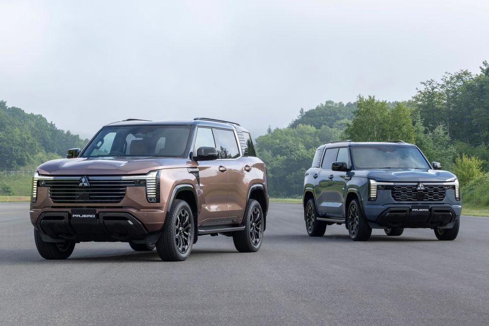 Two Mitsubishi Pajero SUVs — one in brown and one in dark grey — parked on an open area with trees in the background, both seen from the front and side.