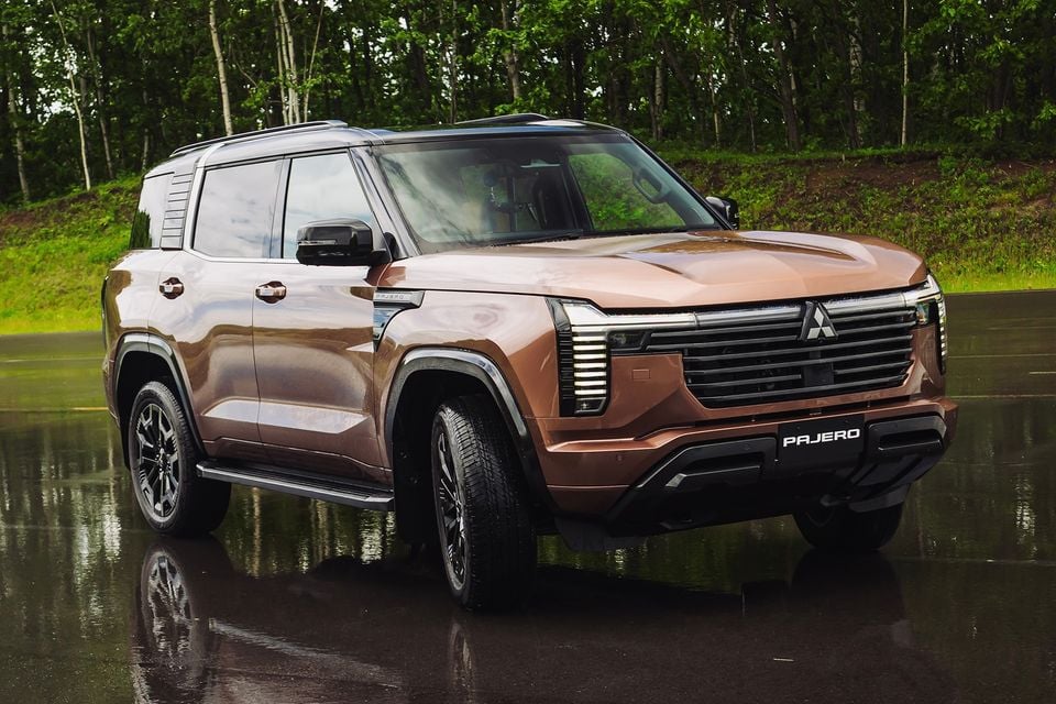 A bronze 2027 Mitsubishi Pajero SUV seen from the front-side angle on a wet surface with trees in the background