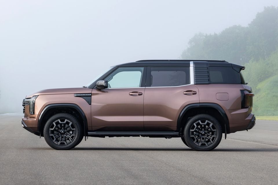 A bronze 2027 Mitsubishi Pajero large SUV seen from the side, parked on a road with misty hills in the background.